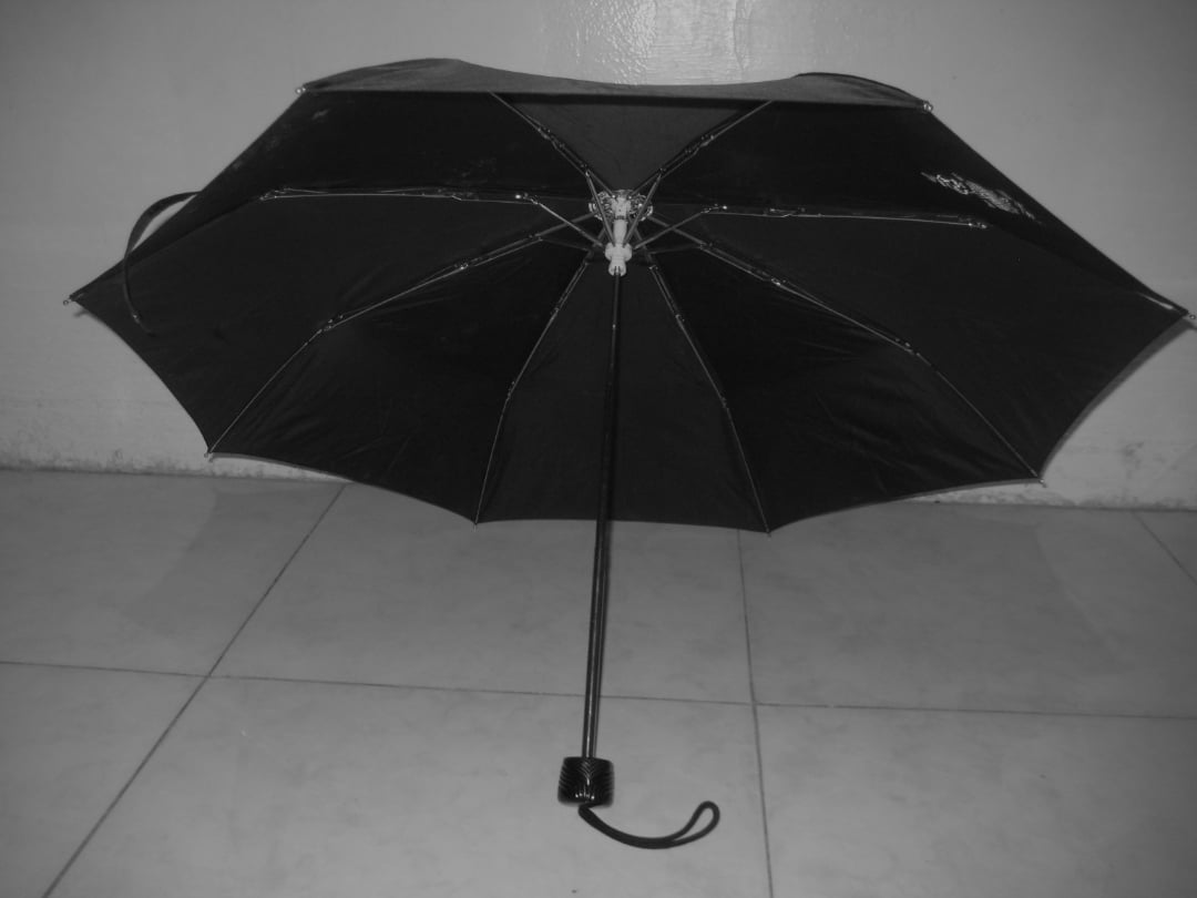 A black umbrella is open and placed upside down on a tiled floor against a plain wall. The umbrella’s handle and shaft are visible in the foreground. The image is in black and white.