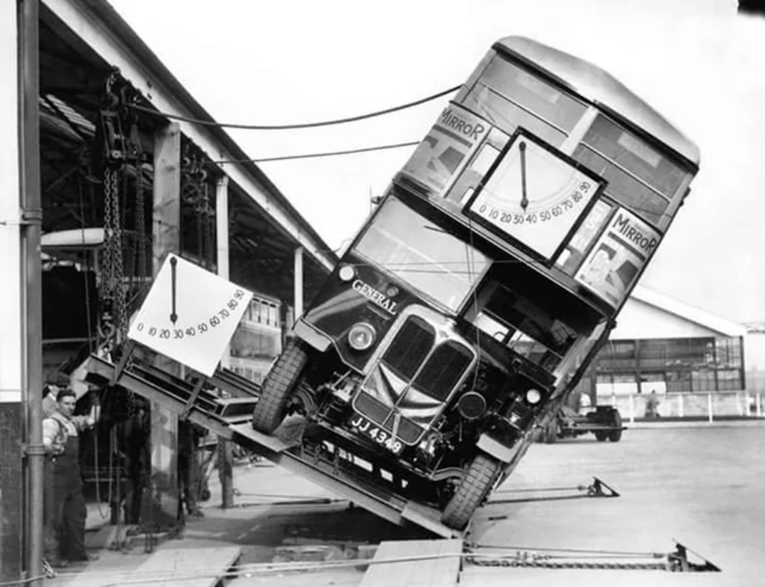 A vintage double-decker bus tilts at a sharp angle on a testing ramp outside a garage, with workers nearby and large dials measuring the tilt.