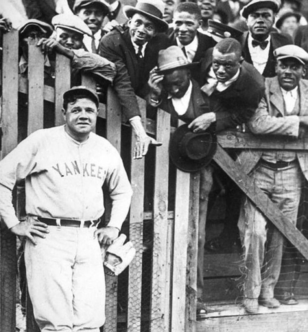 A baseball player in a Yankees uniform stands by a wooden fence, while a group of smiling, well-dressed men lean over the fence to see him. The player poses with hands on hips as the crowd eagerly looks on.