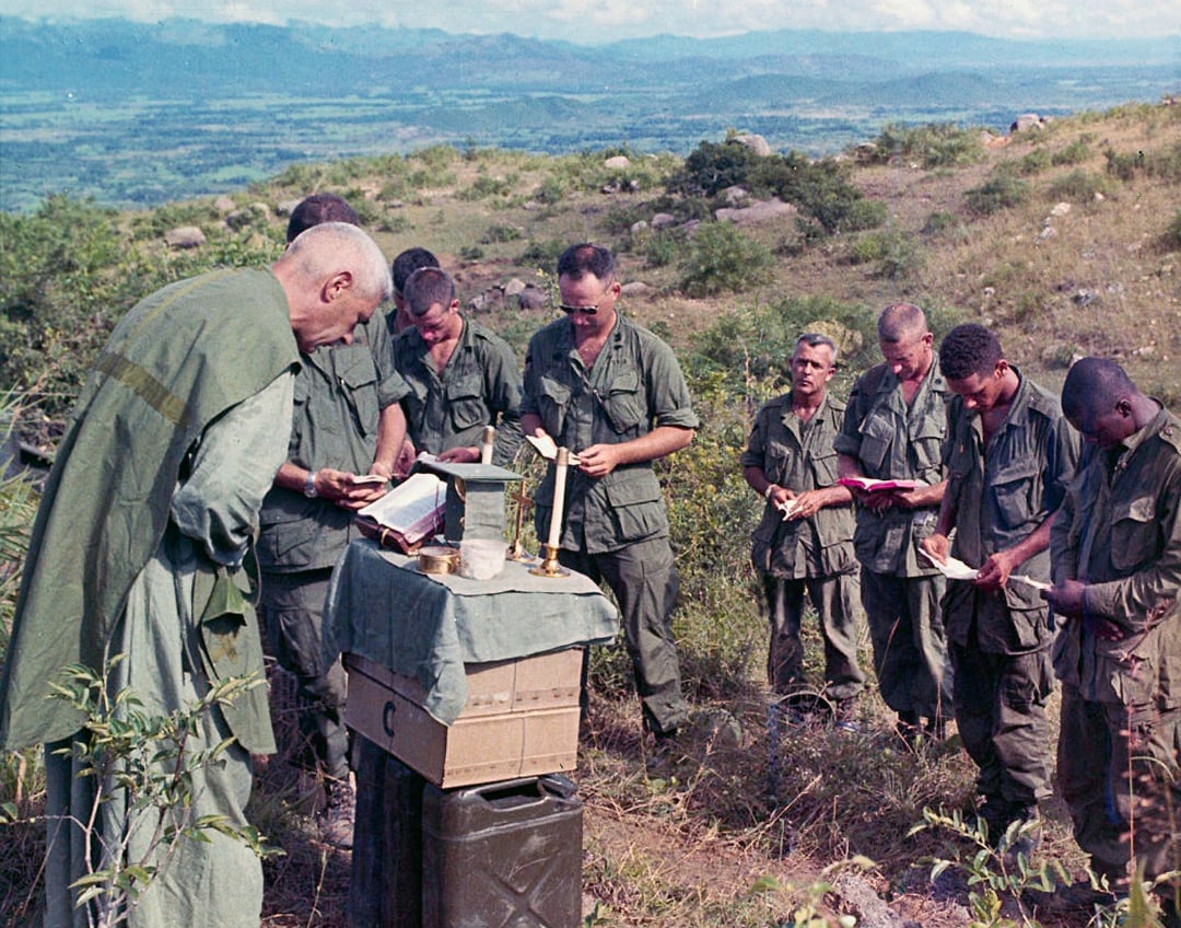 A group of soldiers in uniform stand outdoors as a chaplain conducts a field service. The chaplain stands behind a makeshift altar with candles and a book; the soldiers bow their heads in prayer on a grassy hillside.