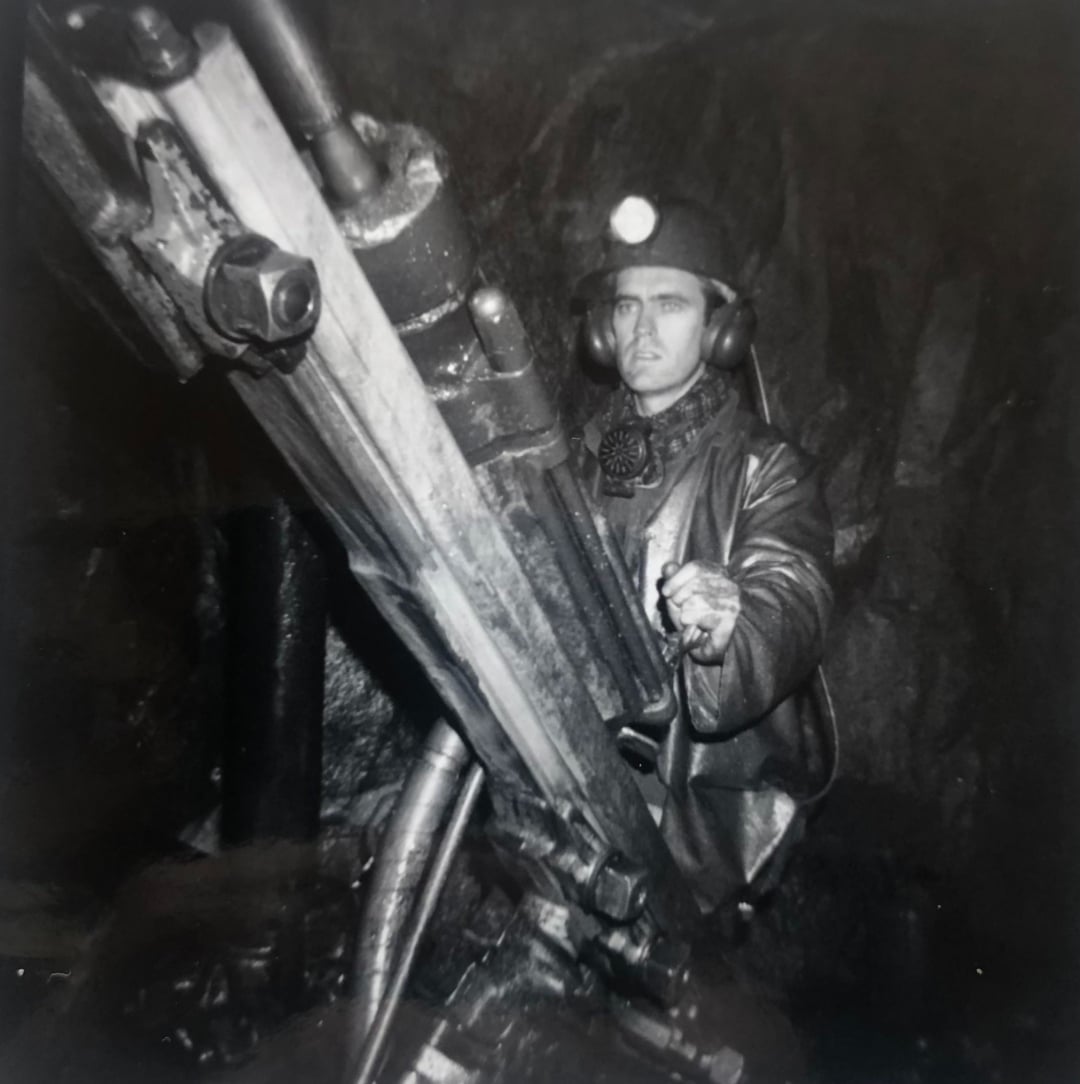 A miner wearing a helmet and headlamp operates heavy drilling machinery underground in a dark, rocky environment.