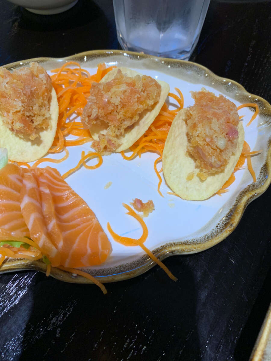 A decorative plate with three crispy chips topped with a raw fish mixture, shredded carrots, and several slices of raw salmon sashimi arranged beside them. A glass of water is in the background.