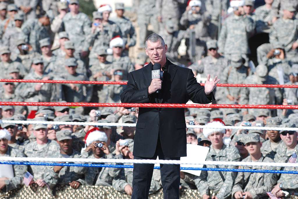 A man in a black suit speaks into a microphone in a wrestling ring, surrounded by ropes, with a large crowd of uniformed soldiers in the background, some wearing Santa hats.