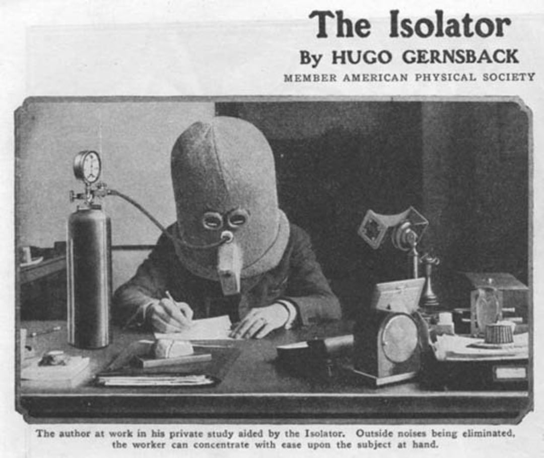 A person wearing a large, helmet-like device called "The Isolator" sits at a desk writing, surrounded by scientific equipment, while an oxygen tank is connected to the helmet.