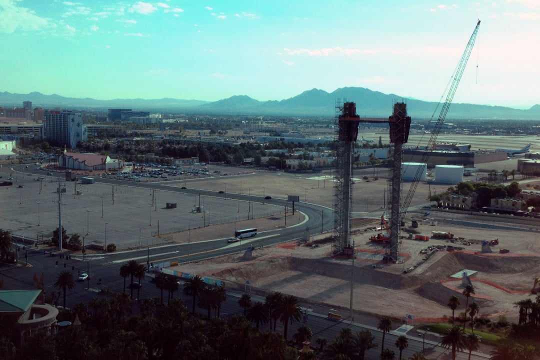 View of a construction site with two tall towers and a crane, surrounded by empty lots and roads, set against a backdrop of city buildings, mountains, and a partly cloudy sky.