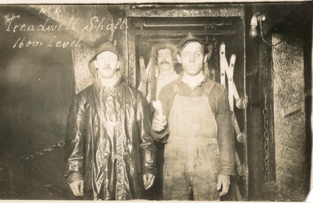Three miners stand in front of a large metal door inside a mine shaft labeled "Treadwell Shaft 1600 Level." Two men stand in the foreground, one holding a candle; a third man stands in the background.