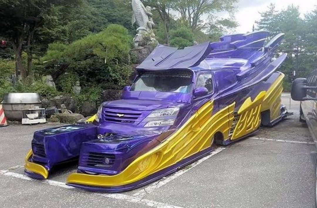 A highly customized van with dramatic purple and yellow bodywork, featuring exaggerated, angular panels and extended fenders, parked in a lot surrounded by trees and statues.