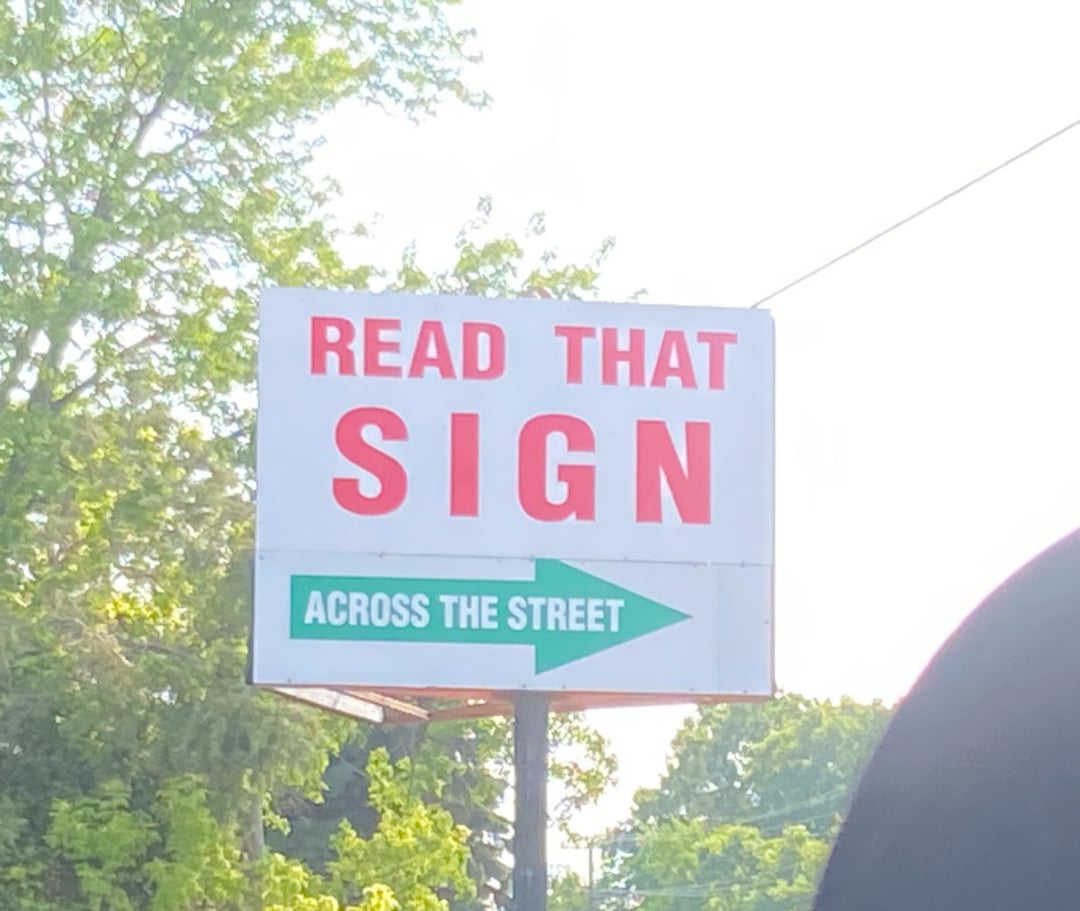 A roadside sign reads "READ THAT SIGN" in red letters, with a green arrow below pointing right and the words "ACROSS THE STREET." Trees and a clear sky are visible in the background.