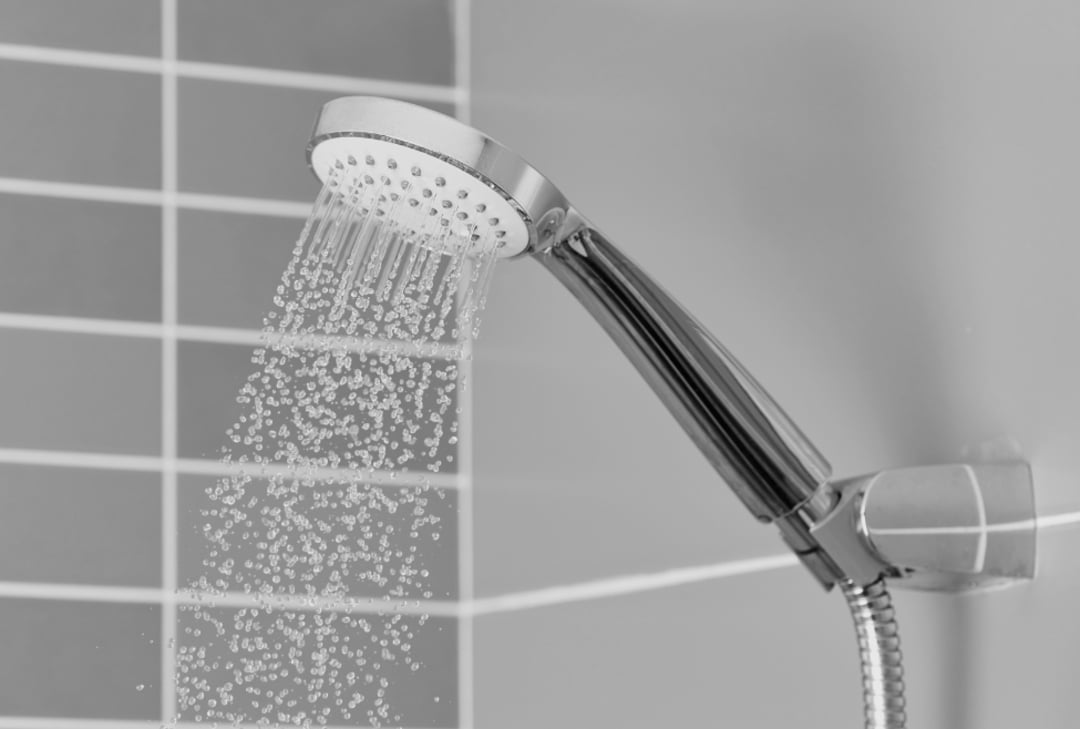 A handheld showerhead attached to a tiled wall, spraying water at an angle. The photo is in black and white.