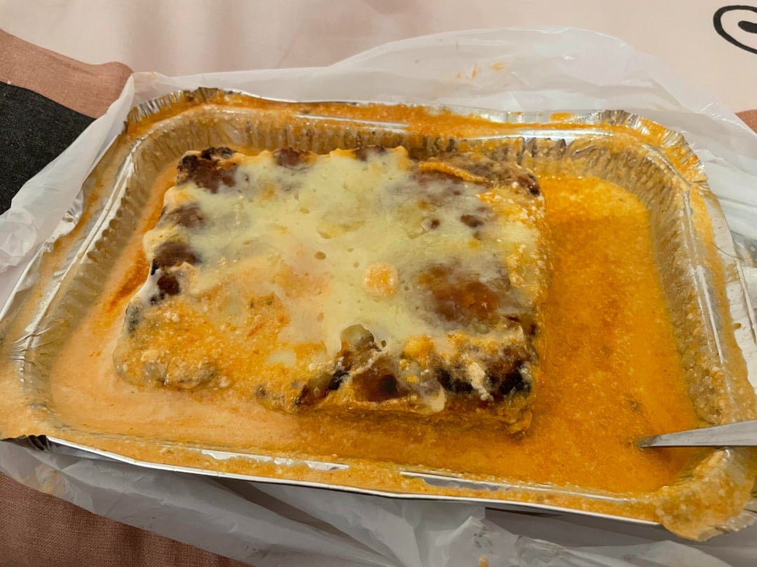 A rectangular foil tray filled with a serving of cheesy baked lasagna in an orange sauce, with a spoon resting on the side. The tray sits on a plastic sheet and a tablecloth.