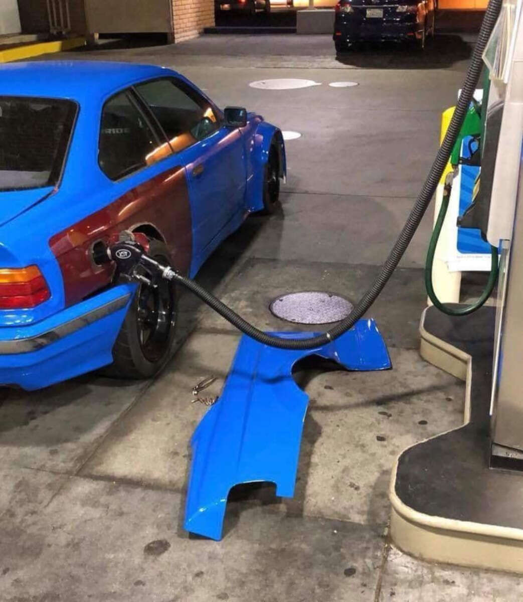 A blue car at a gas station with its rear fender detached and lying on the ground; the fuel nozzle is inserted into the fender instead of the car’s gas tank.
