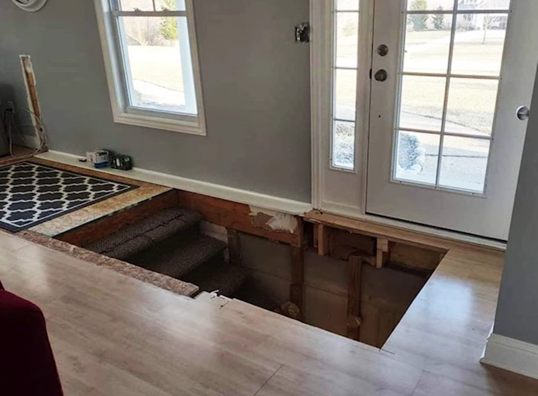 A living room with light wood floors and gray walls has an exposed staircase hole near glass doors, showing unfinished stairs and missing floorboards, creating a hazard.