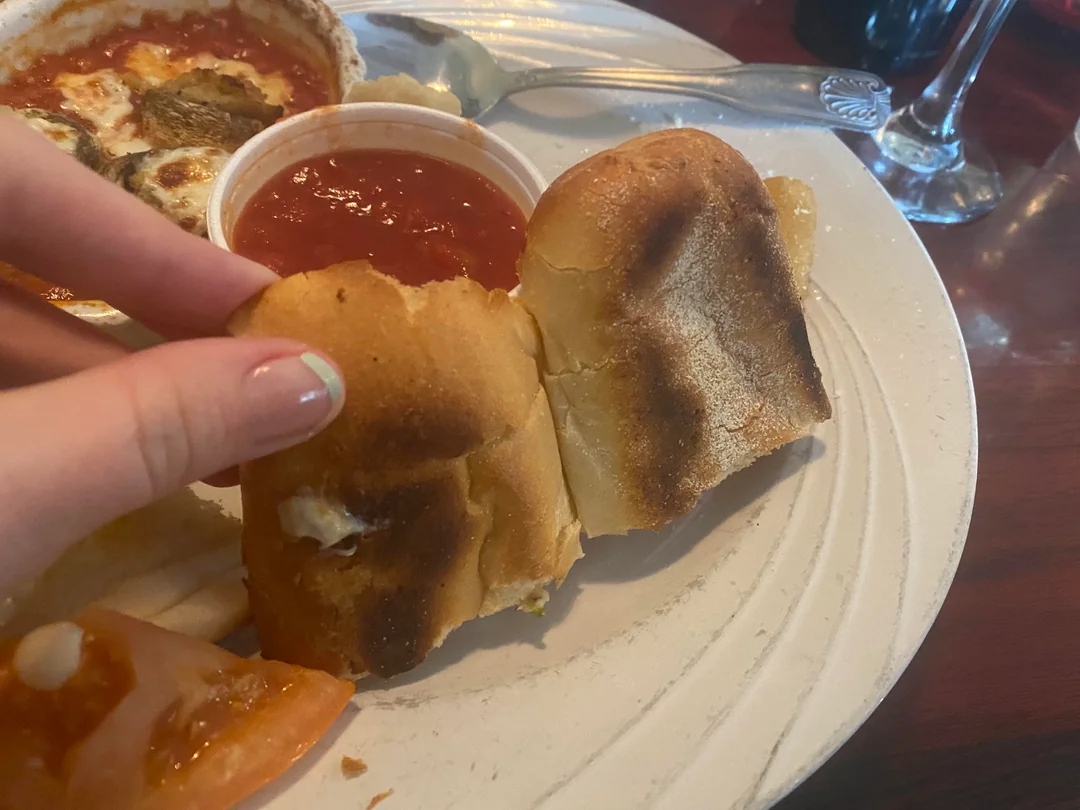 A hand holding a piece of toasted bread next to another piece on a white plate, with a cup of red marinara sauce and part of a slice of tomato visible. A partially eaten dish and a spoon are also in the background.