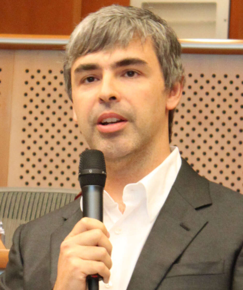 A man with short gray hair, wearing a white shirt and dark suit jacket, speaks into a handheld microphone in an indoor setting with a perforated wood panel background.