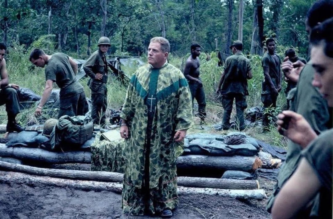 A military chaplain in a camouflage robe with a cross necklace stands among soldiers in a jungle clearing, with troops preparing equipment and resting in the background.