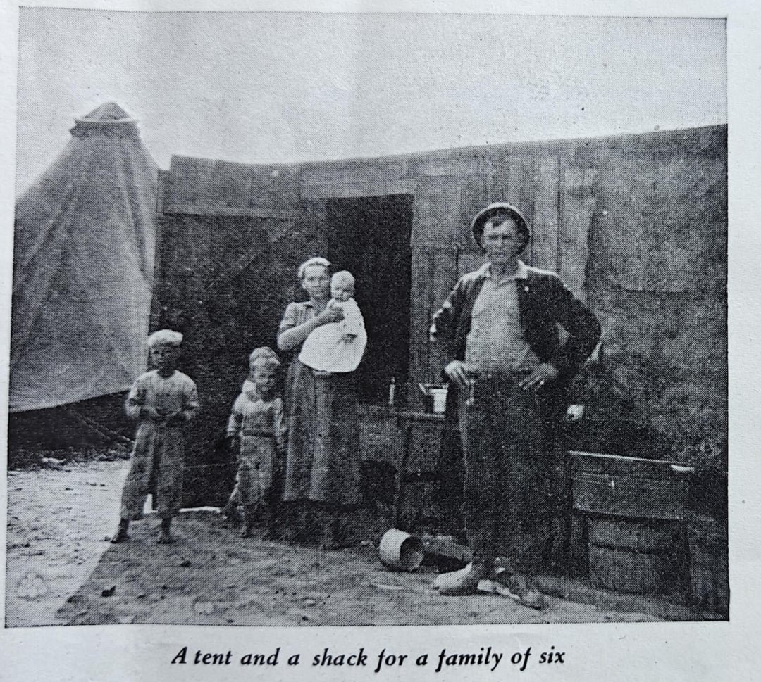 A black and white photo of a family of six, including two adults and four children, standing outside a small shack with a tent in the background. The adults and children wear simple clothing.