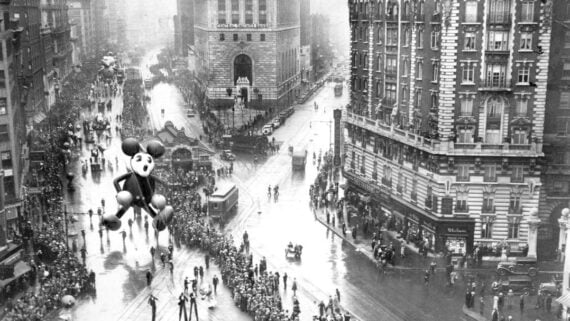 Black-and-white photo of a parade with a large Mickey Mouse balloon, marching bands, and crowds lining wet city streets surrounded by tall buildings. 1930s