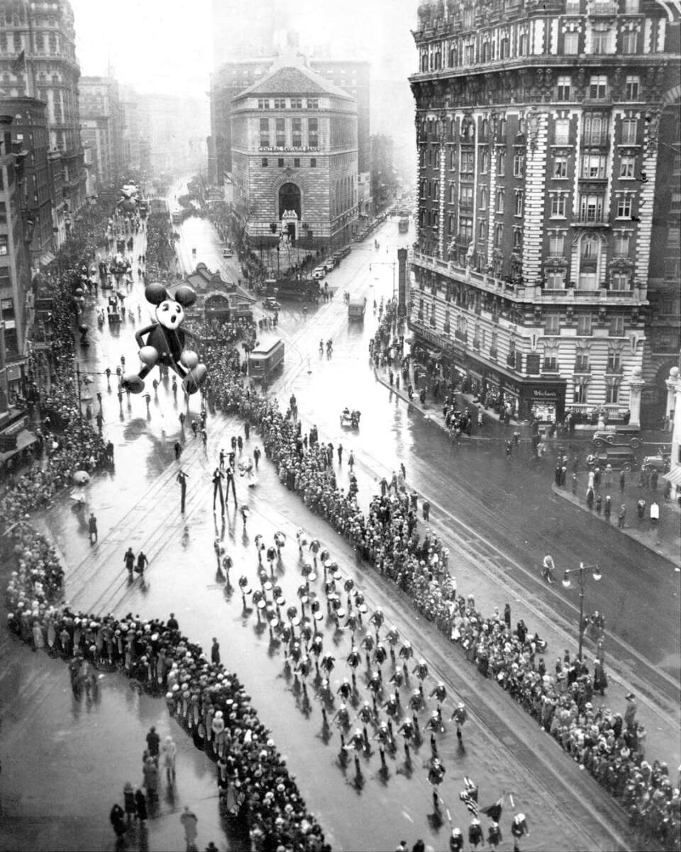 Black and white photo of a crowded city street during a parade, featuring a large Mickey Mouse balloon, marching bands, and spectators lining the sidewalks; buildings and wet pavement visible.