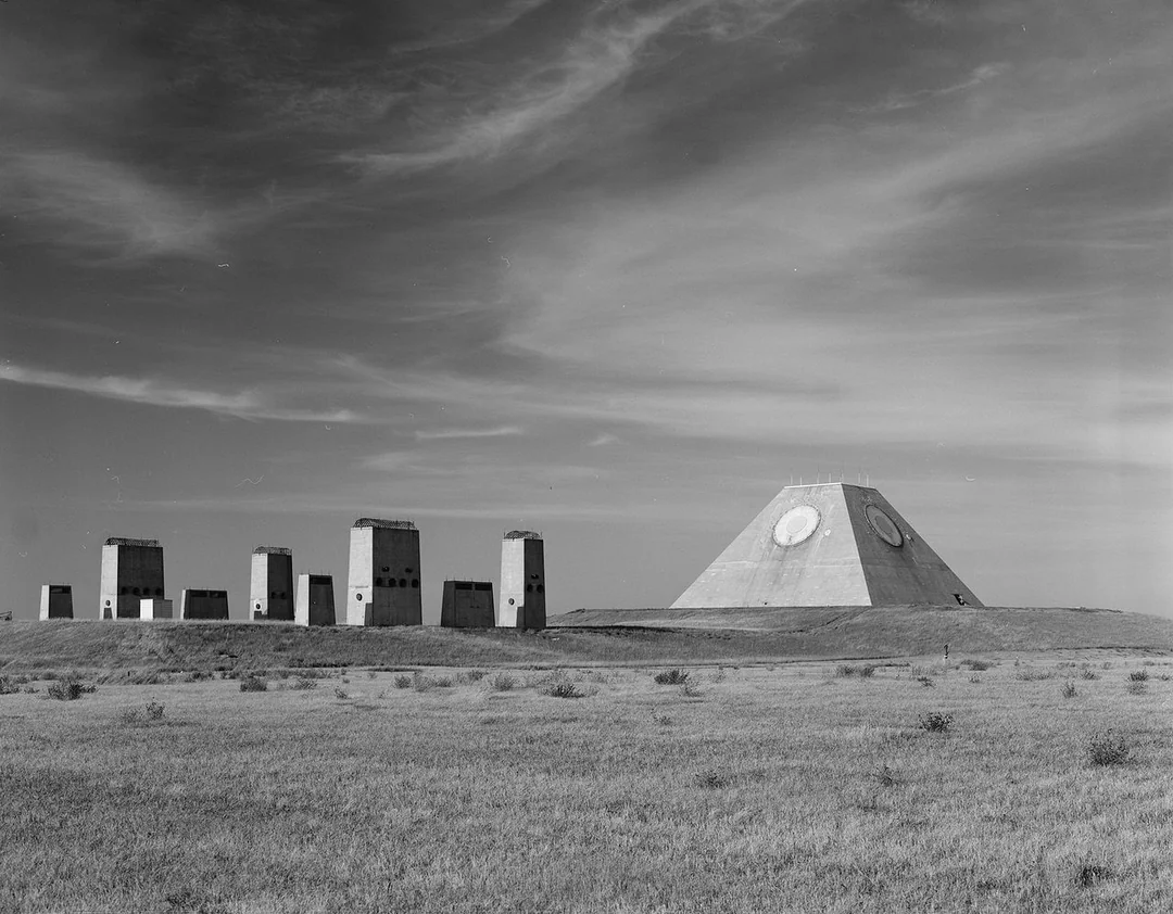 A large concrete pyramid with circular markings stands in a flat, grassy landscape, surrounded by several tall, rectangular structures under a partly cloudy sky.