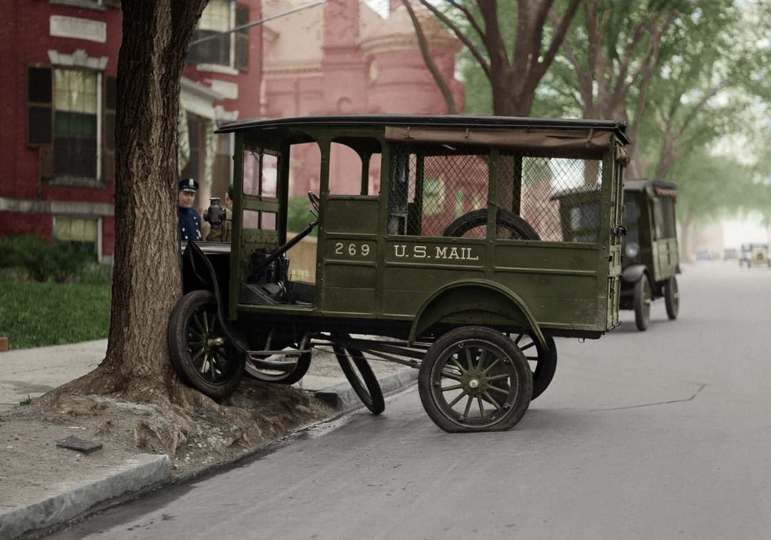 A vintage green U.S. Mail truck has crashed into a tree on a city street. A uniformed mail carrier stands nearby. Another similar vehicle is parked further down the road. The scene is set in an early 20th-century neighborhood.