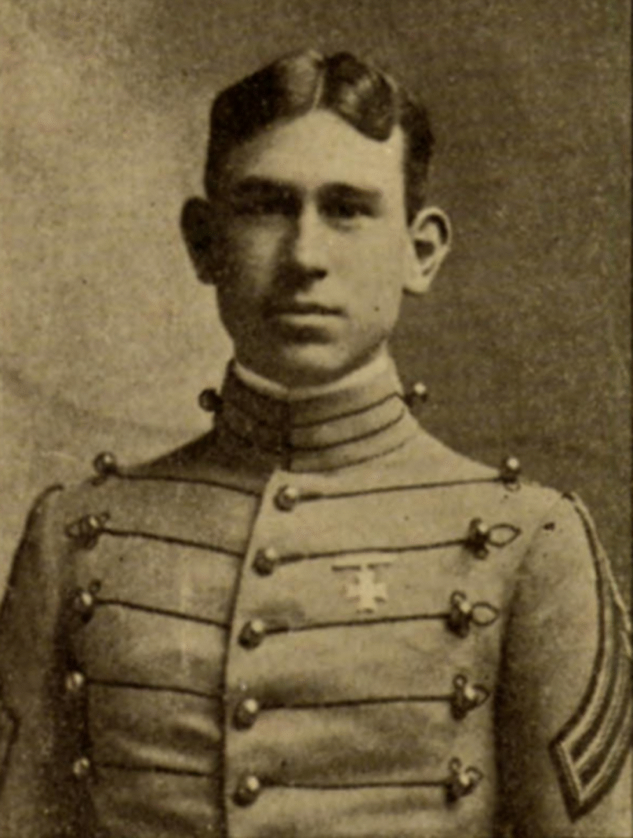 A young man in an old-fashioned military uniform with decorative buttons poses for a portrait. The photo is sepia-toned and appears to be from the early 20th century.