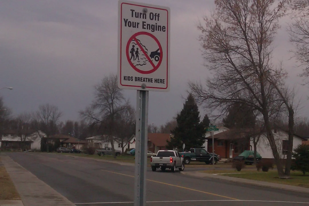 A street sign reads "Turn Off Your Engine. Kids Breathe Here" with a crossed-out image of a running car near children. Suburban houses and leafless trees are visible in the background.