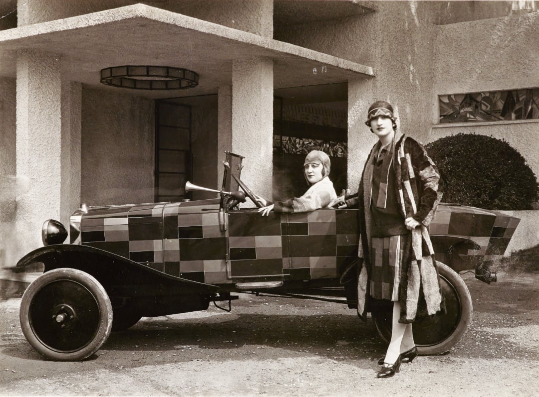 Two women pose with a vintage car featuring a bold, blocky checkered pattern; one sits behind the wheel while the other stands beside the car, both wearing 1920s-style clothing, in front of a textured building.