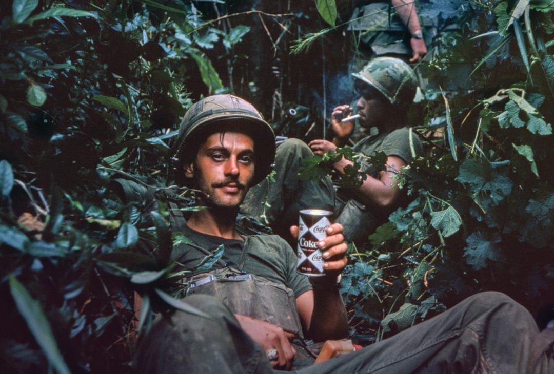 Two soldiers in military uniforms sit among dense jungle foliage. The man in front holds a can of Coca-Cola and looks at the camera, while the man behind smokes a cigarette. Both appear tired but relaxed.
