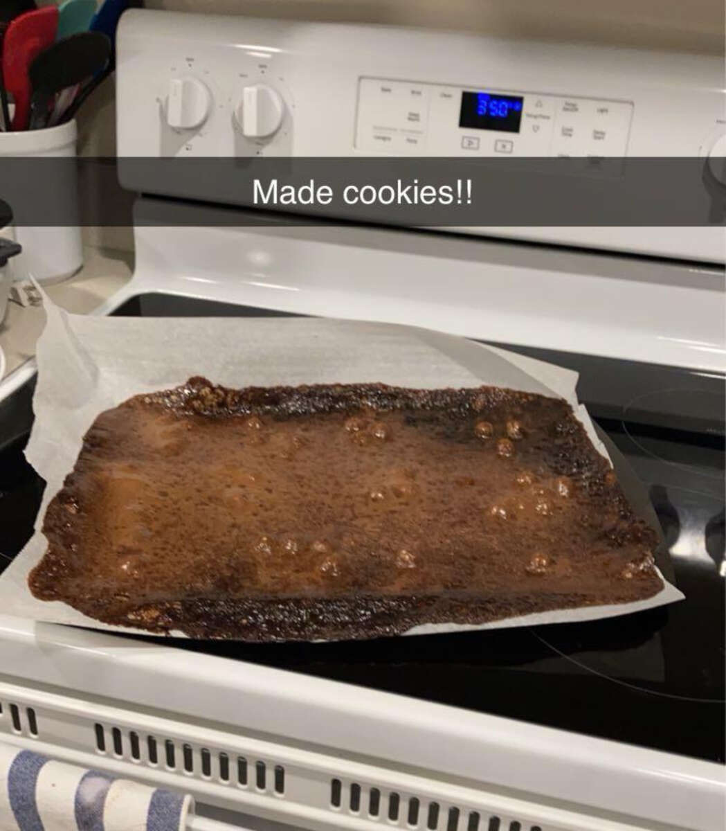 A baking tray covered with parchment paper shows a large, flat, burnt sheet of cookie dough with uneven spots, resting on a stove. A caption above says, “Made cookies!!”