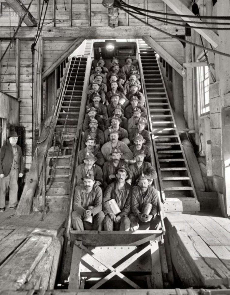 A group of coal miners sit in rows on a mine car, posed for a photo inside a wooden mining facility, with stairs on both sides and industrial equipment visible overhead.