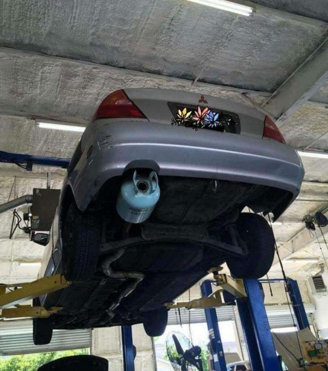 A silver car is elevated on a hydraulic lift inside a garage. An LPG gas cylinder is humorously attached where the muffler should be, and the license plate is covered with flower stickers.