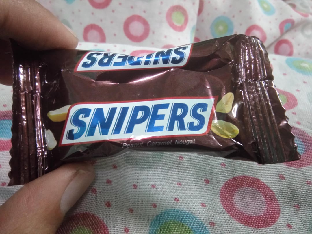 A hand holding a small, brown candy wrapper labeled "SNIPERS" with images of peanuts; the background is white fabric with colorful rings and polka dots.