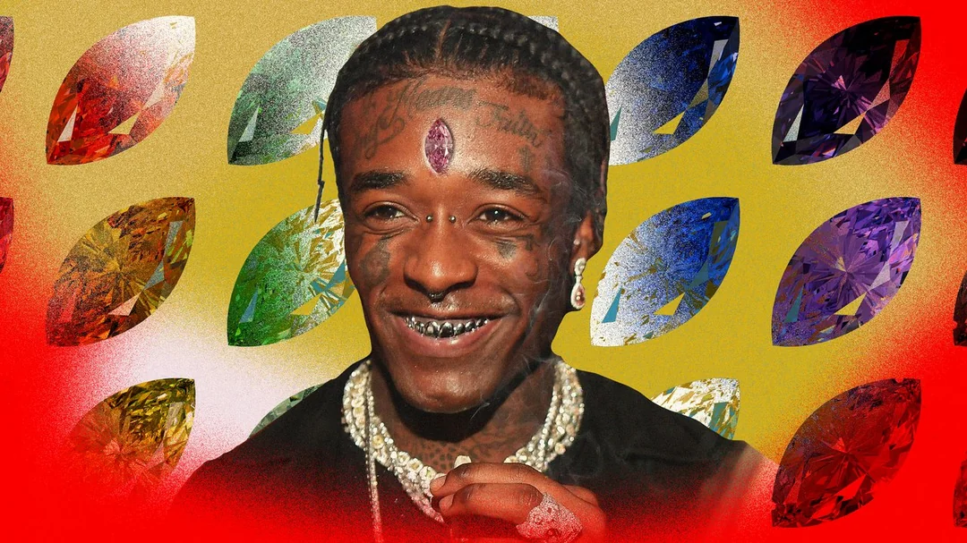 A smiling person with facial tattoos, braids, facial piercings, and a large pink gem embedded in their forehead stands in front of a colorful, patterned background featuring various gemstones.