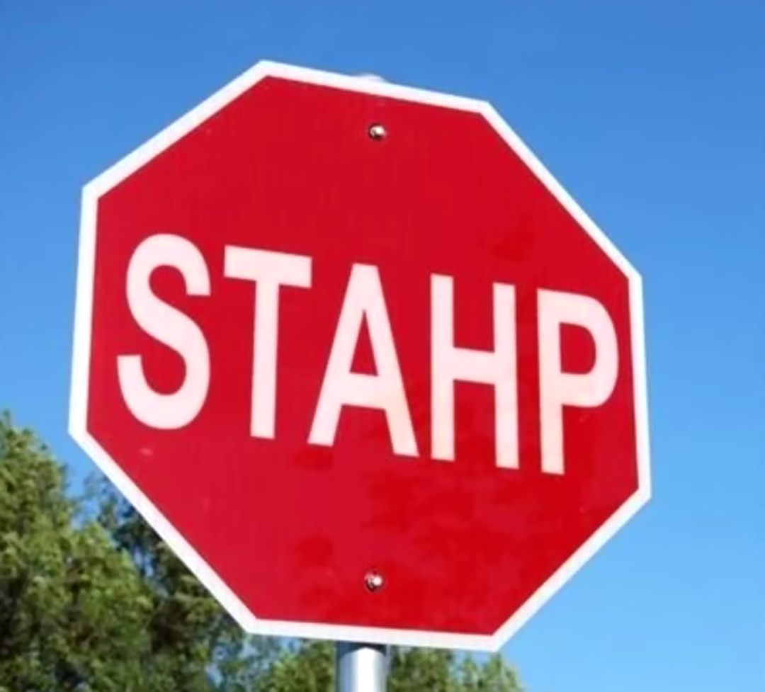A red octagonal street sign with the word “STAHP” written in white letters instead of “STOP,” against a clear blue sky and some green trees.