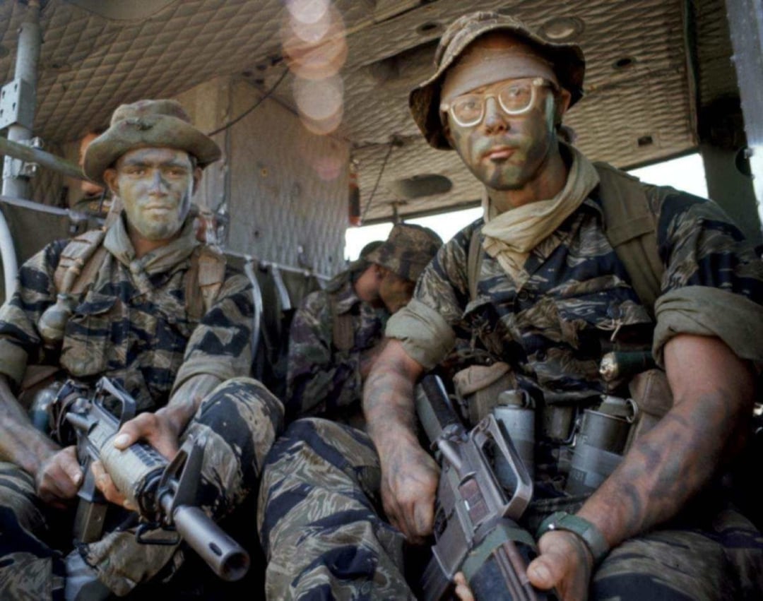 Two soldiers in camouflage face paint and tiger stripe uniforms sit inside a military helicopter, holding rifles and gear, with two more similarly dressed soldiers visible in the background.