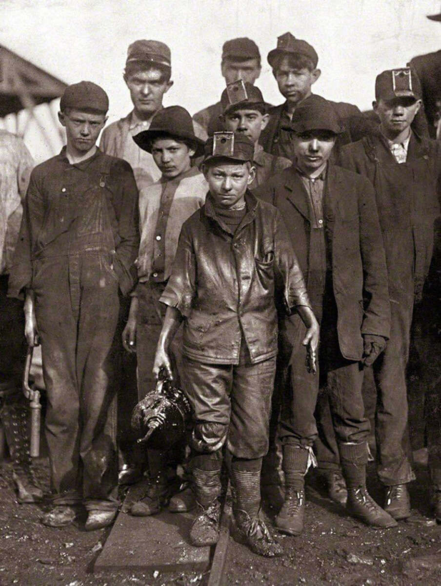 A group of young boys wearing work clothes and caps stand together in a coal mine setting, some holding tools and lanterns. Their faces and clothes appear dirty, showing evidence of hard labor.