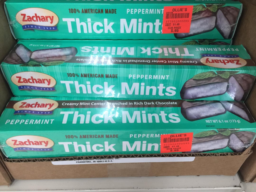 Three packages of Zachary brand Thick Mints peppermint candies, covered in dark chocolate, are stacked in a display box. Each package has a red sale sticker from Ollie's Bargain Outlet.