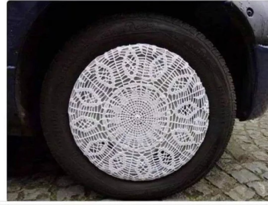 A car tire with a white crocheted doily-style cover over the wheel, parked on a cobblestone surface.