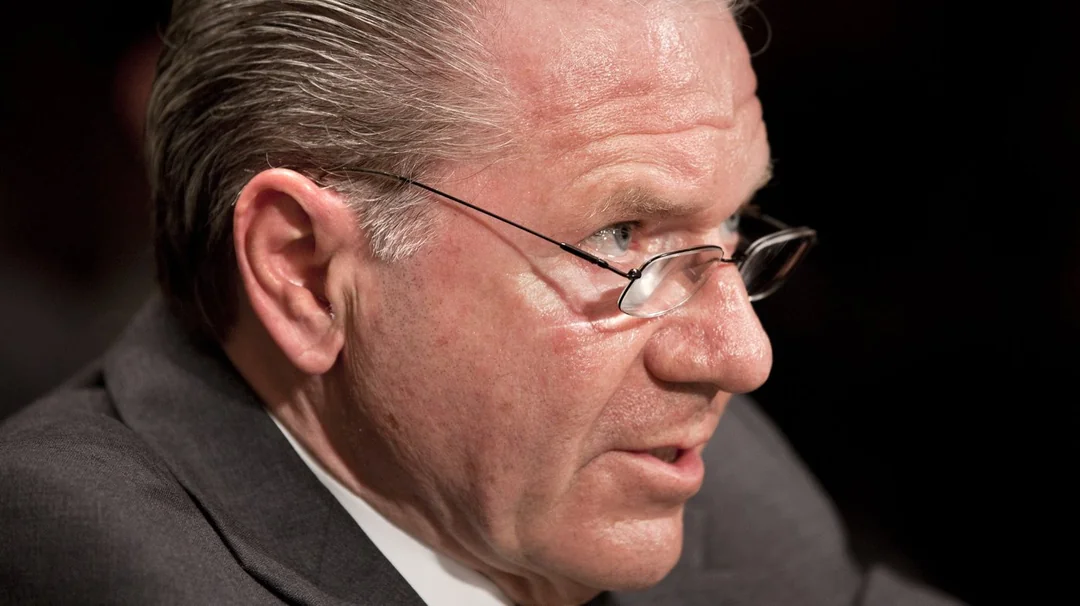 A middle-aged man with gray hair and glasses, wearing a dark suit, is shown in close-up, speaking or listening intently, with a serious expression and a blurred dark background.