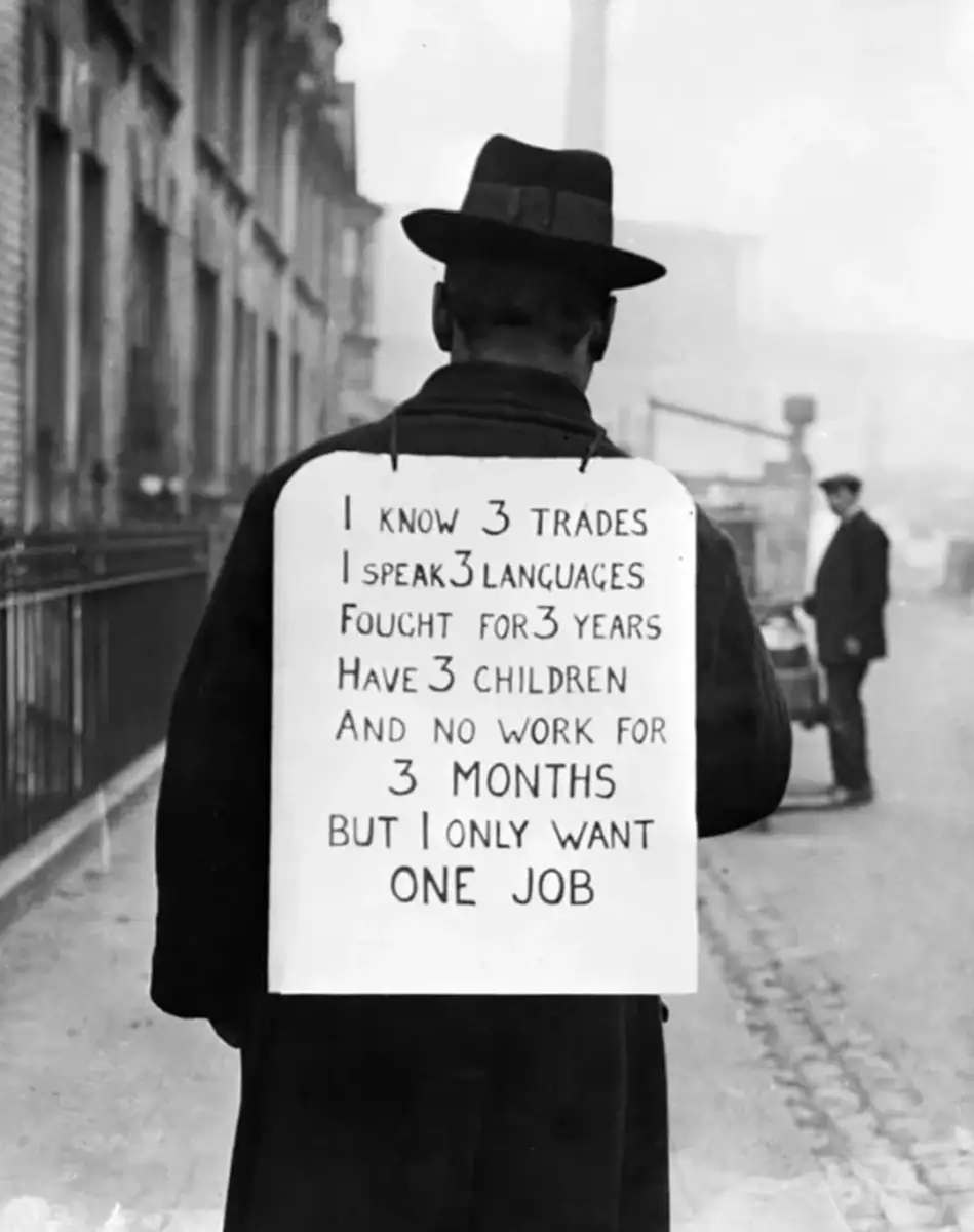 A man in a hat and coat stands on a sidewalk, viewed from behind. He wears a sign reading, "I know 3 trades, I speak 3 languages, fought for 3 years, have 3 children, and no work for 3 months—but I only want one job.