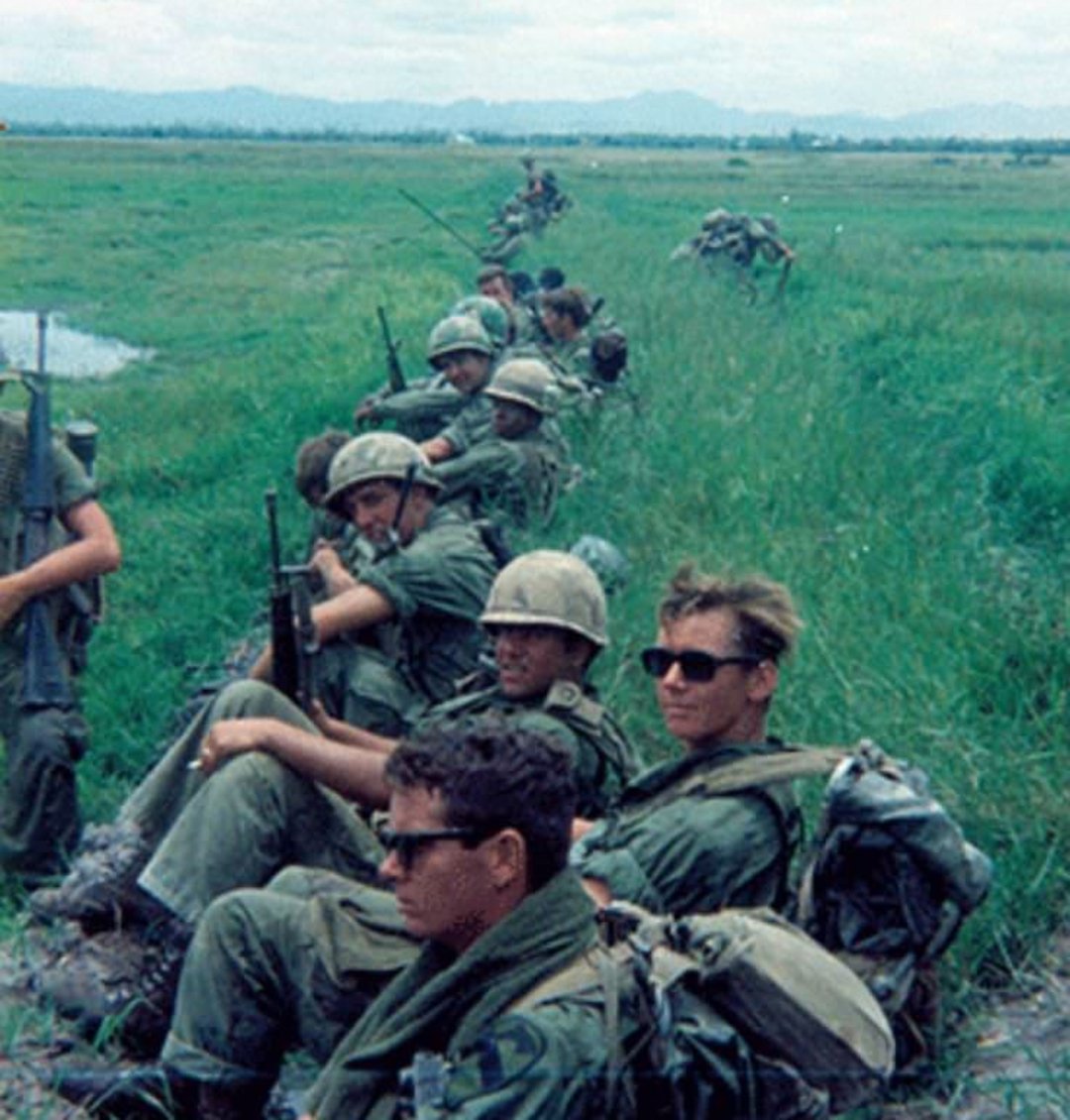 A group of soldiers in green uniforms and helmets sit and rest in tall grass in a field, forming a long line, with gear and rifles beside them. The landscape is open with distant mountains under a cloudy sky.