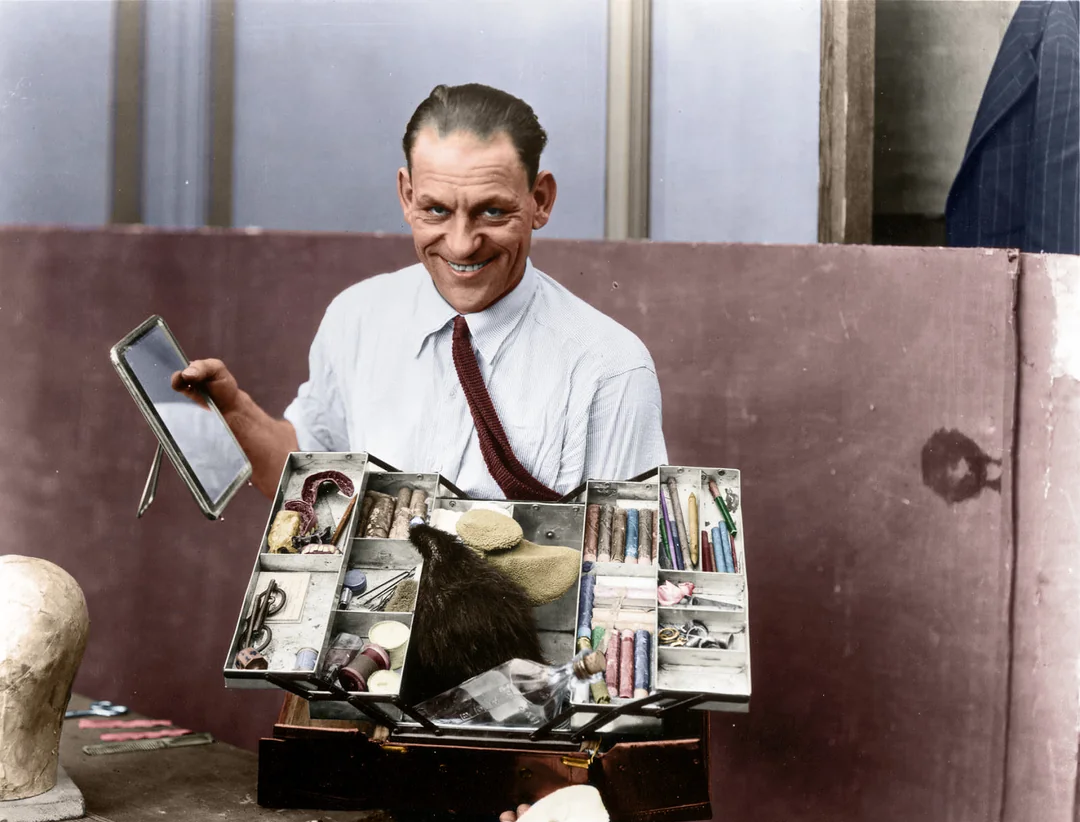 A smiling man in a shirt and tie holds a small mirror and open makeup case filled with brushes, powders, and other tools, standing beside a mannequin head.