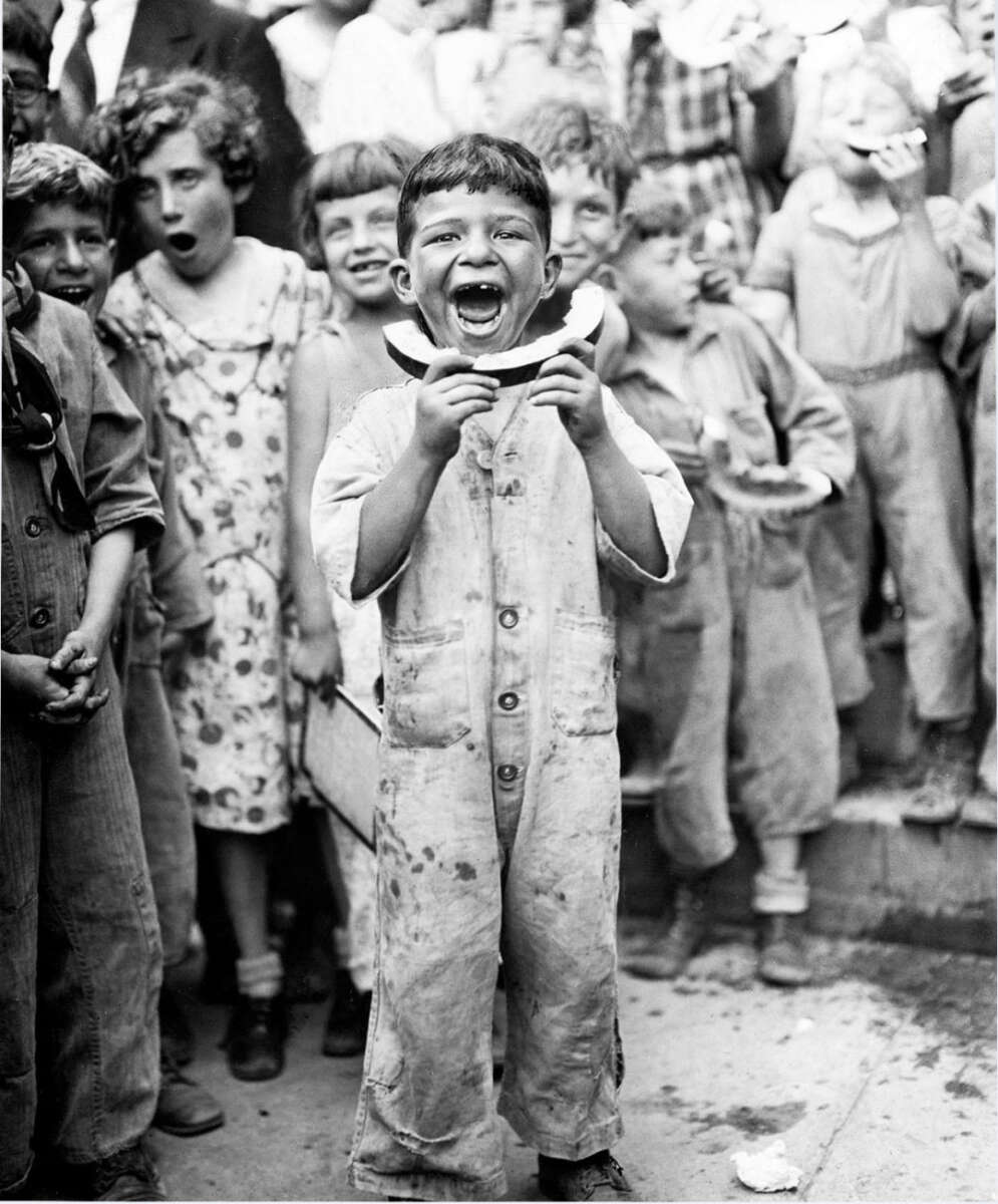 A young boy in overalls stands smiling and holding a watermelon rind, surrounded by other children who are also eating watermelon and laughing during what appears to be a joyful summer gathering.