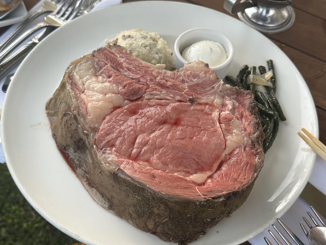A plate with a thick slice of rare prime rib, mashed potatoes, a small cup of horseradish sauce, and green beans, served on a white dish with silverware on a wooden table.