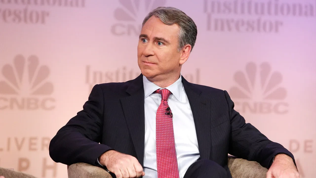 A man in a dark suit, white shirt, and red tie sits on a chair at a business event, looking to the side. The background displays CNBC and Institutional Investor logos.