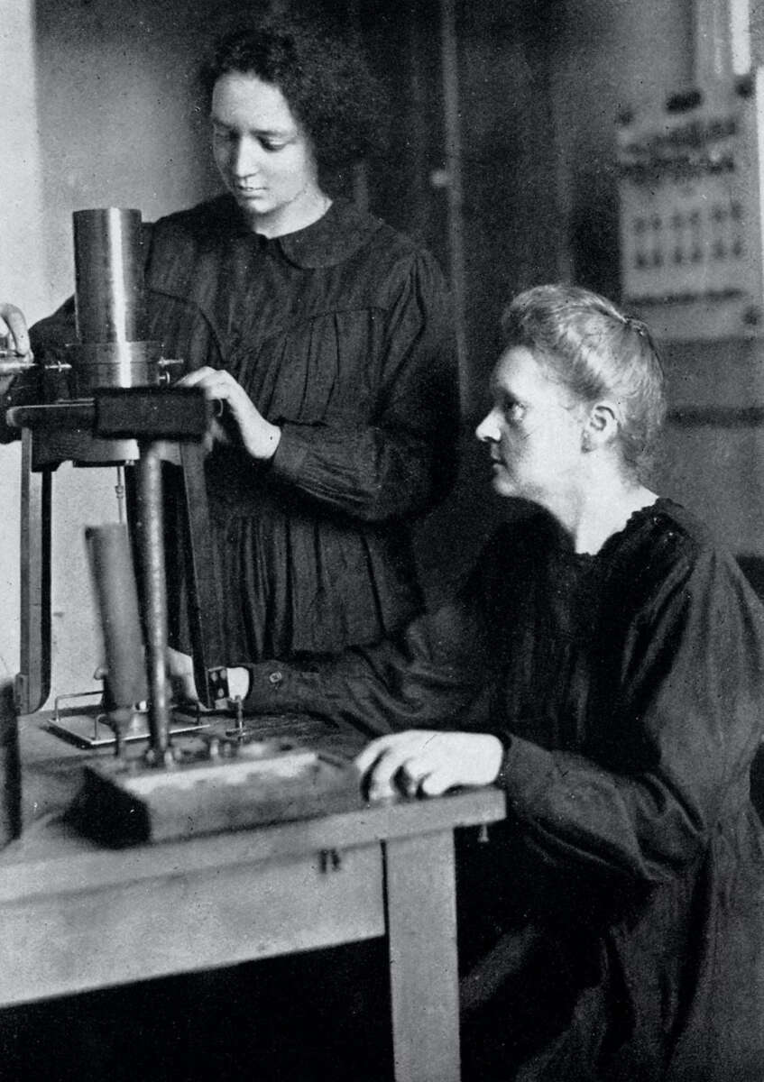 Two women in dark dresses work with scientific equipment in a laboratory. One is seated, focusing on a device, while the other stands beside her, adjusting an instrument on the tabletop apparatus.