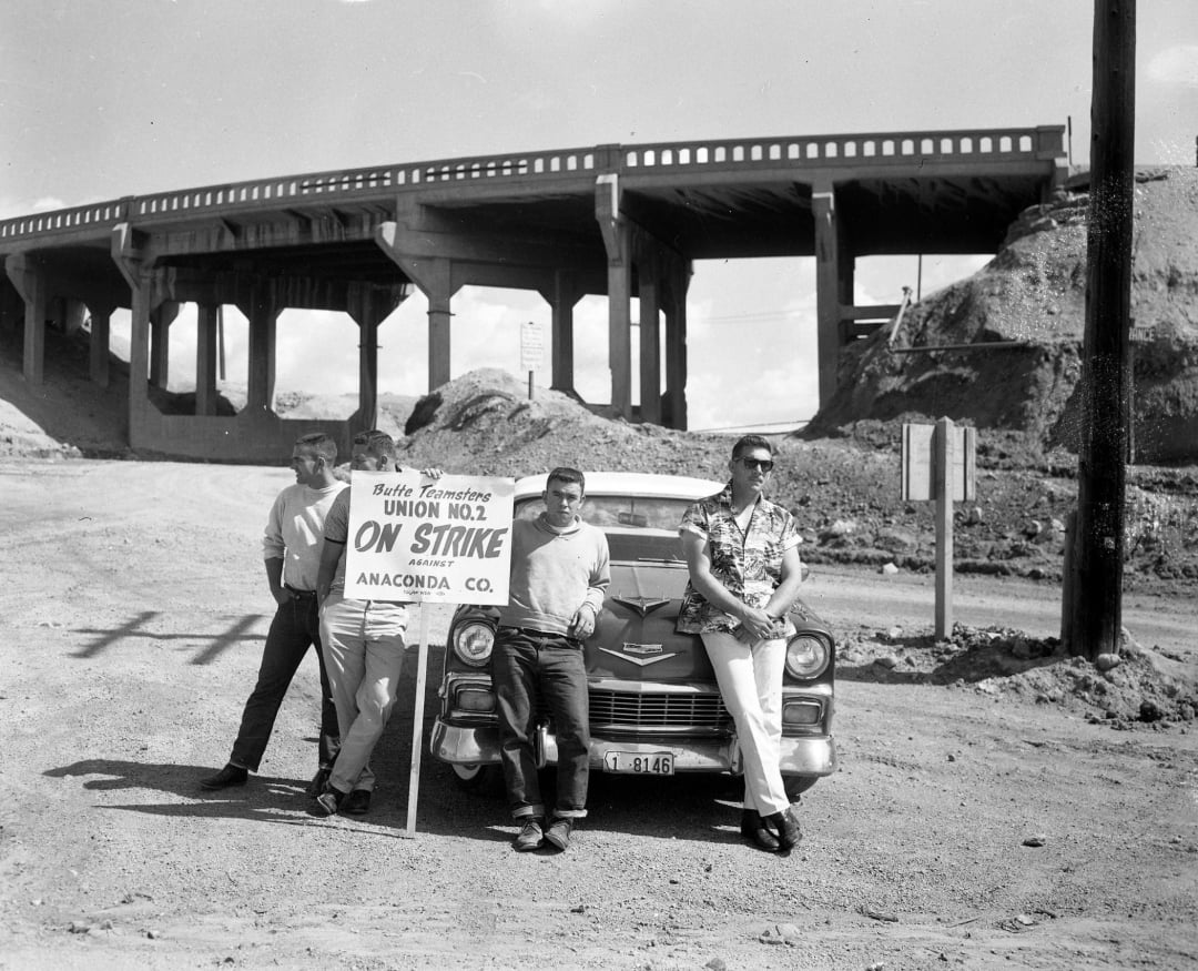 Three men lean against a vintage car, holding a sign that reads "Butte Teamsters Union No. 2 On Strike Anaconda Co." An overpass and dirt mounds appear in the background on a sunny day.