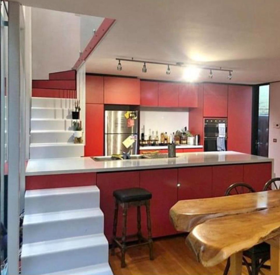 Modern kitchen with red cabinets, a white countertop, a built-in oven, a fridge, and an open bar area; white stairs lead up on the left, and a wooden dining table with chairs is in the foreground.
