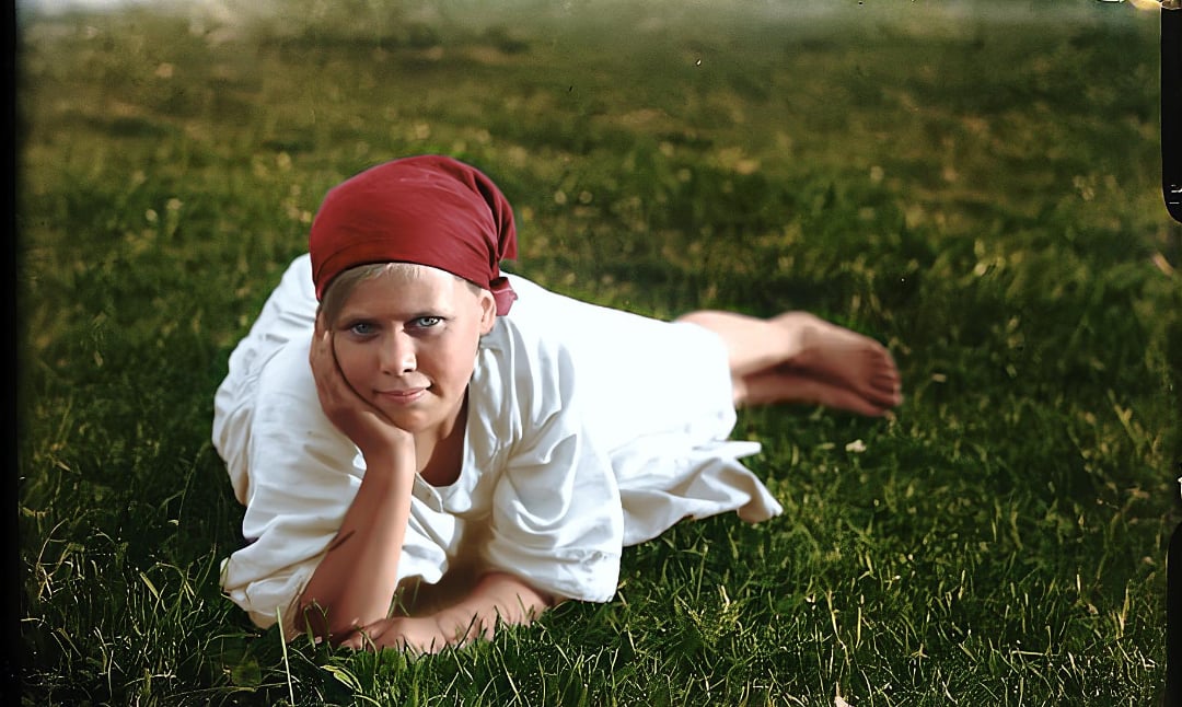 A person wearing a white outfit and a red headscarf lies on green grass, propping their head on one hand and looking at the camera with a calm expression.