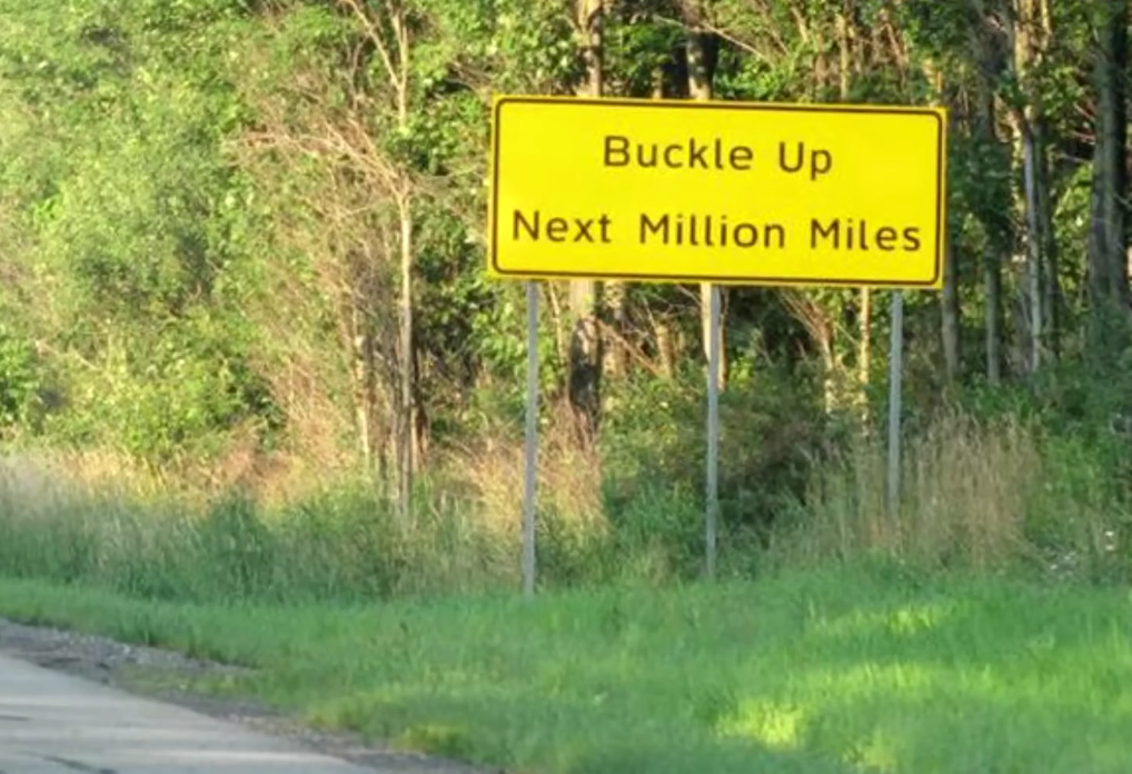 A bright yellow road sign on a grassy roadside reads, "Buckle Up Next Million Miles," with trees and greenery in the background.